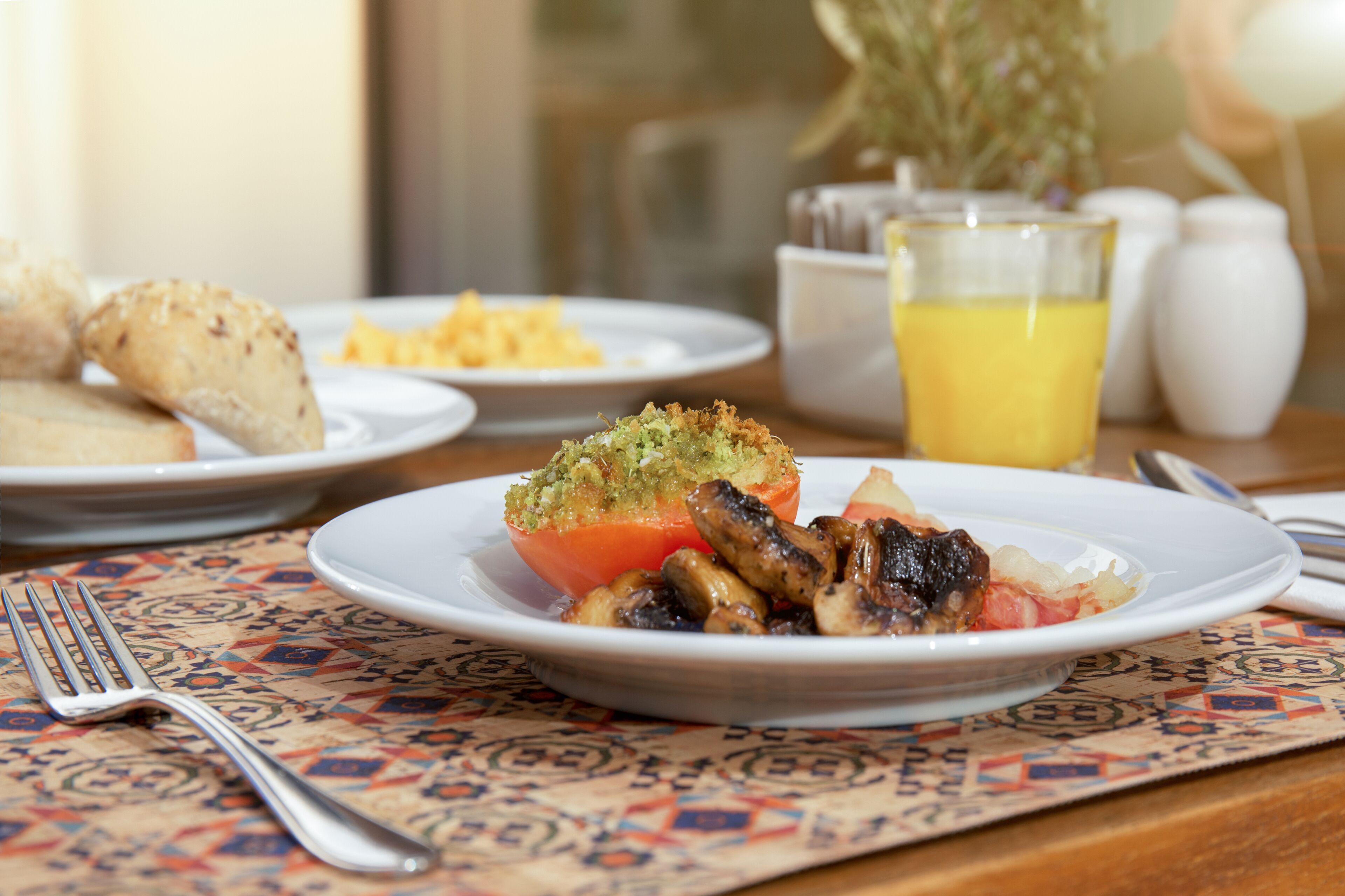 Une table mise pour le petit-déjeuner, avec des assiettes remplies de nourriture, un verre de jus d’orange et une fourchette posée sur une nappe à motifs.