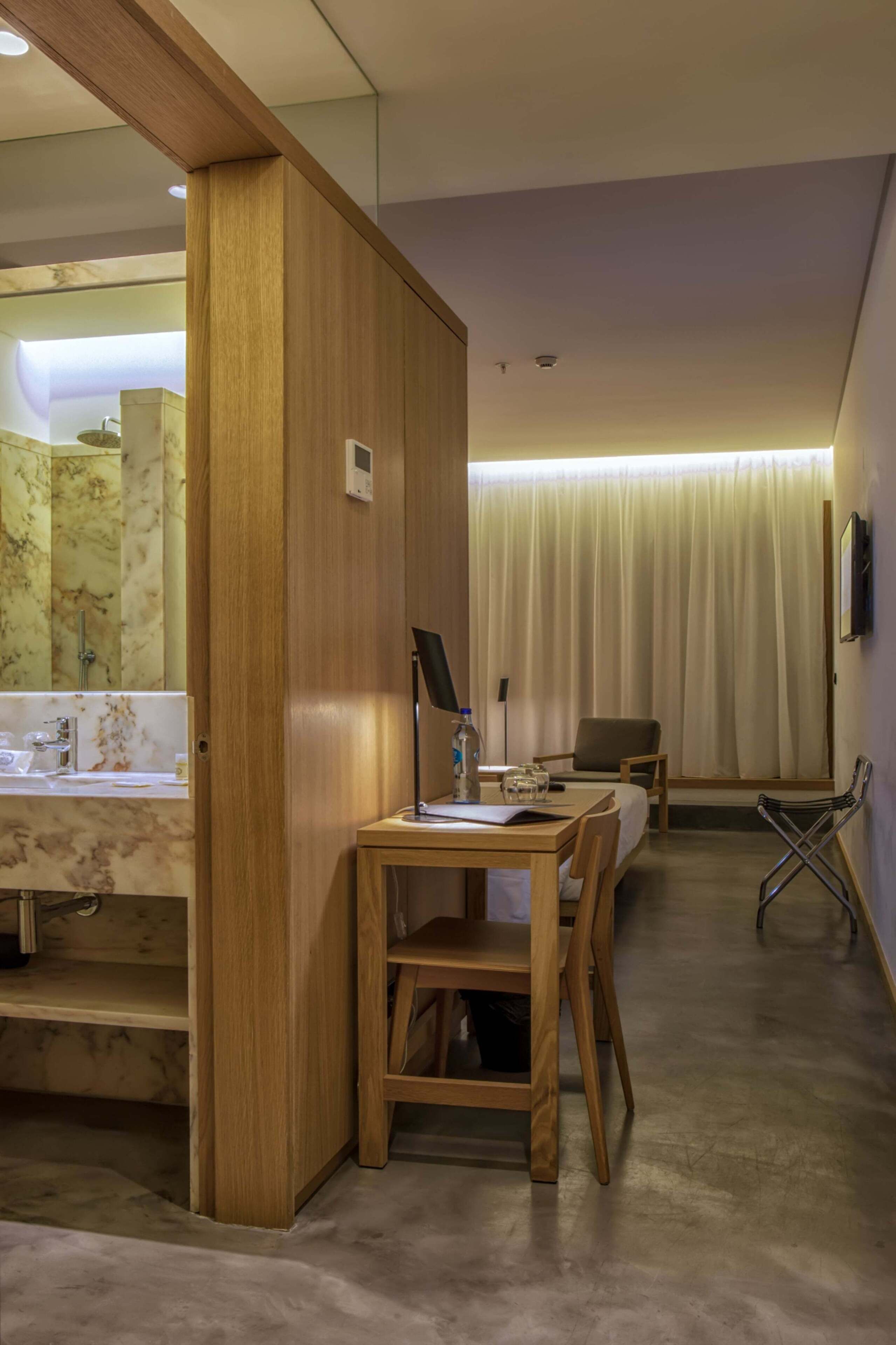 Habitación de hotel con un escritorio de madera, una silla, una cama, un baño con lavamanos de mármol y un ventanal con cortinas.