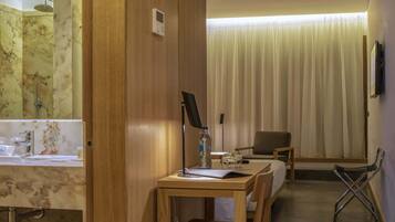 Habitación de hotel con un escritorio de madera, una silla, una cama, un baño con lavamanos de mármol y un ventanal con cortinas.