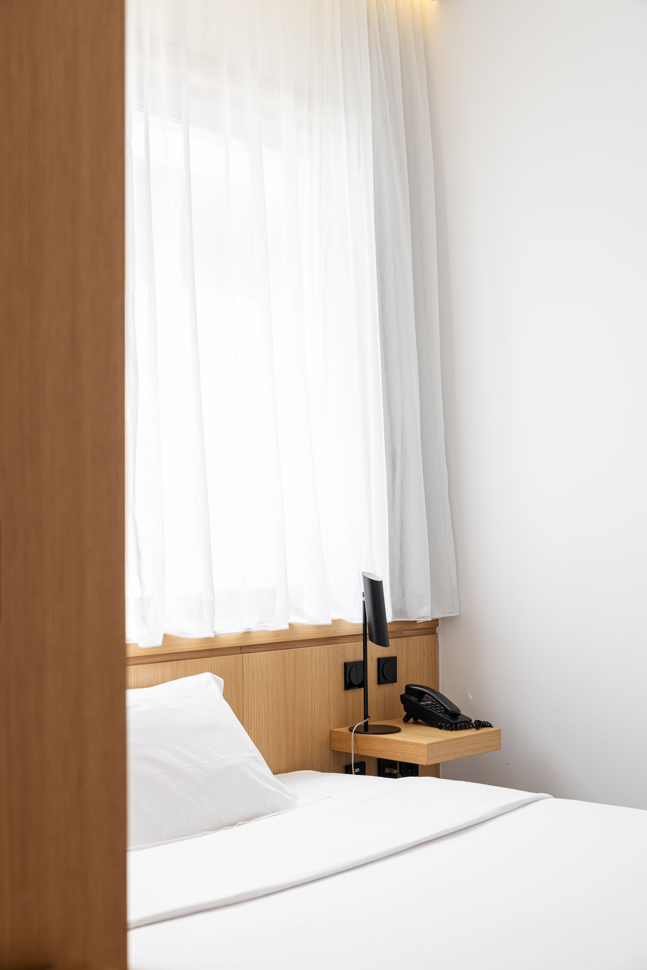 Una habitación de hotel con una cama, una mesita de noche con un teléfono y cortinas translúcidas.