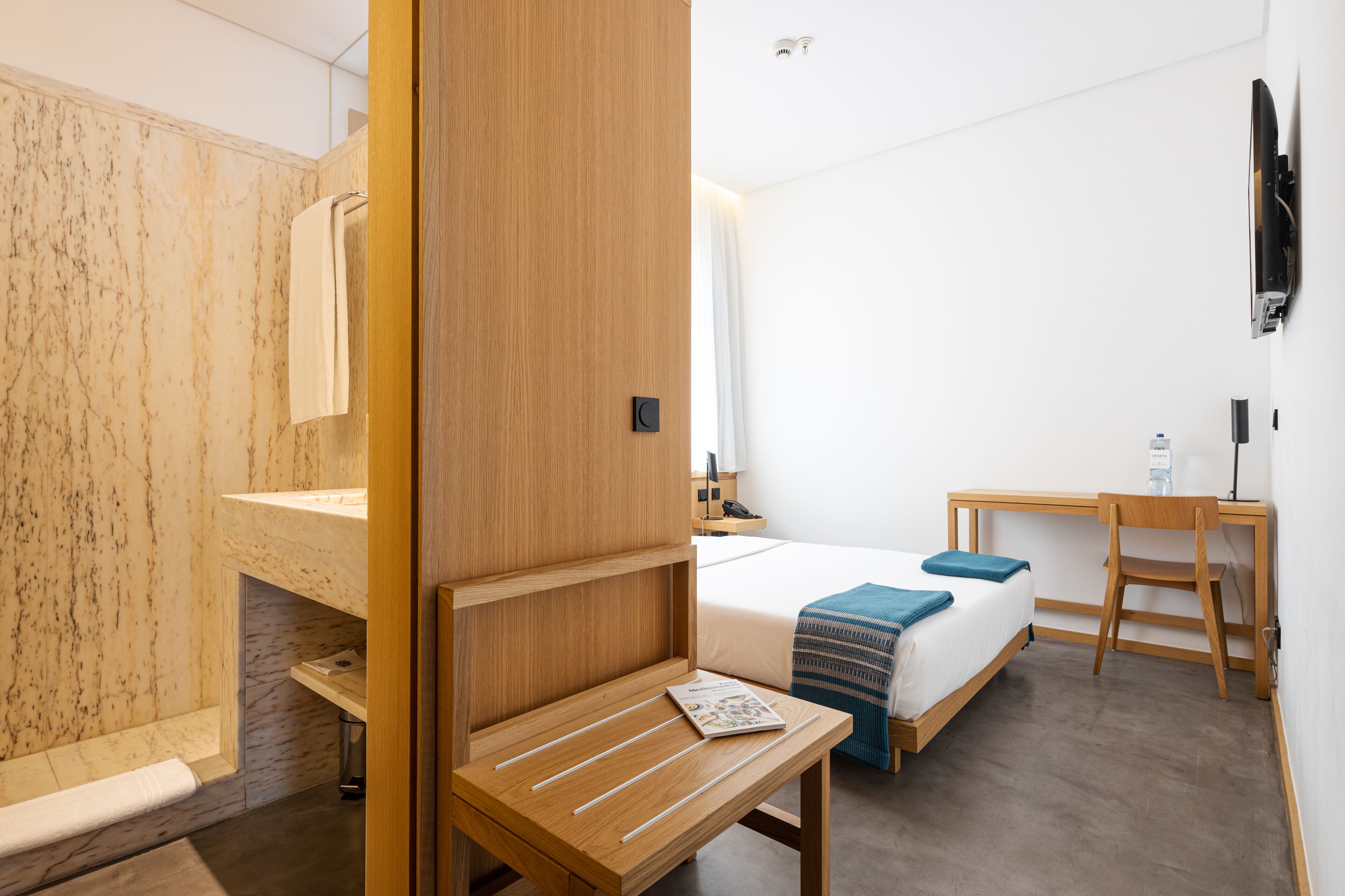 A modern hotel room with a wooden vanity, a bed with a blue blanket, a desk with a chair, and a mounted television.