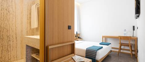 A modern hotel room with a wooden vanity, a bed with a blue blanket, a desk with a chair, and a mounted television.