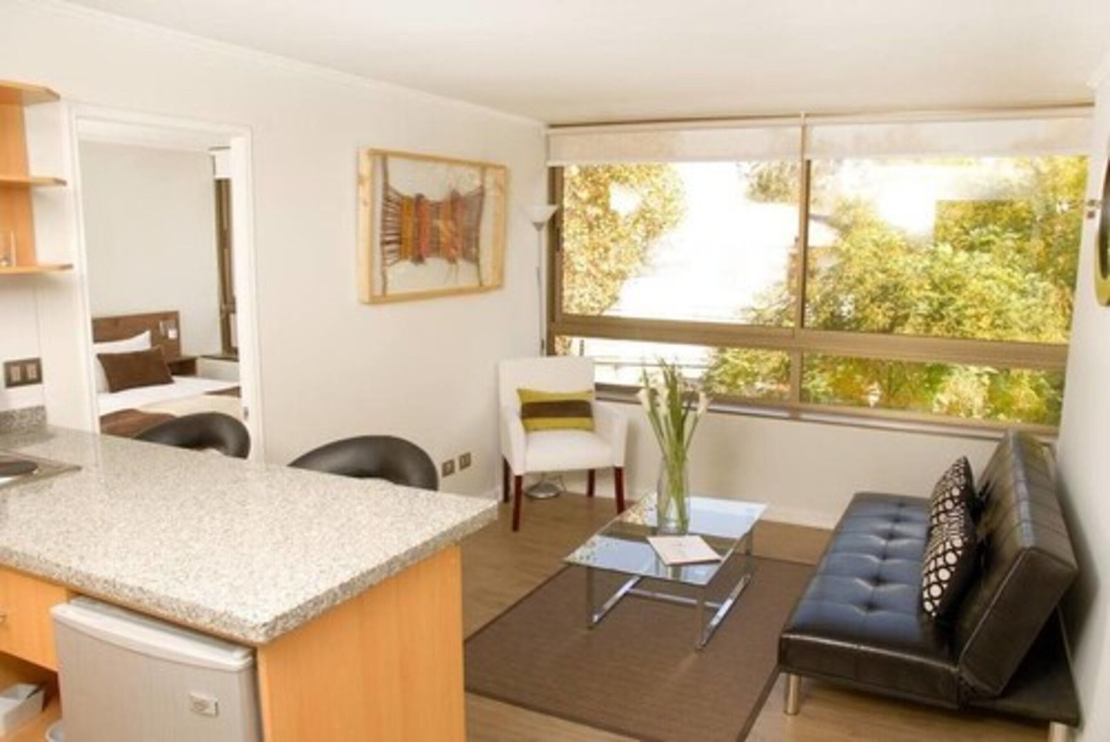 Una sala de estar moderna con área de cocina, un sofá, una mesa de comedor y un ventanal con vista a árboles.