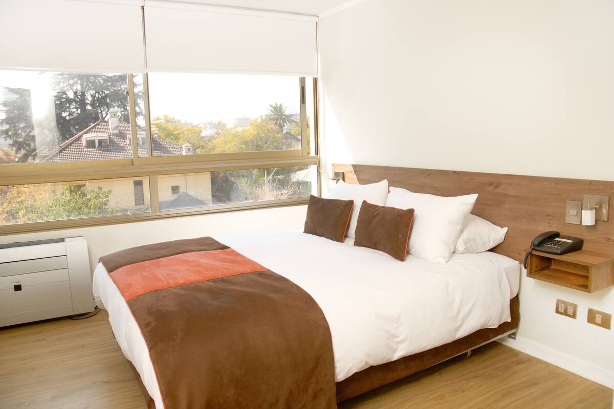 Un dormitorio con una cama grande, un ventanal con vistas a edificios y árboles, una mesita de noche con un teléfono y un calefactor en el suelo.