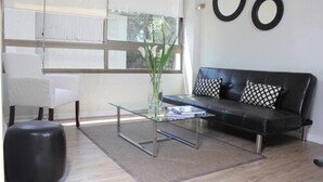 A modern living room with a glass coffee table, a black leather sofa with patterned cushions, a white armchair, and a large window with blinds.