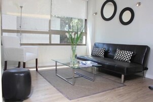 Una sala de estar moderna con una mesa de centro de vidrio, un sofá de cuero negro con cojines estampados, un sillón blanco y un amplio ventanal con persianas.