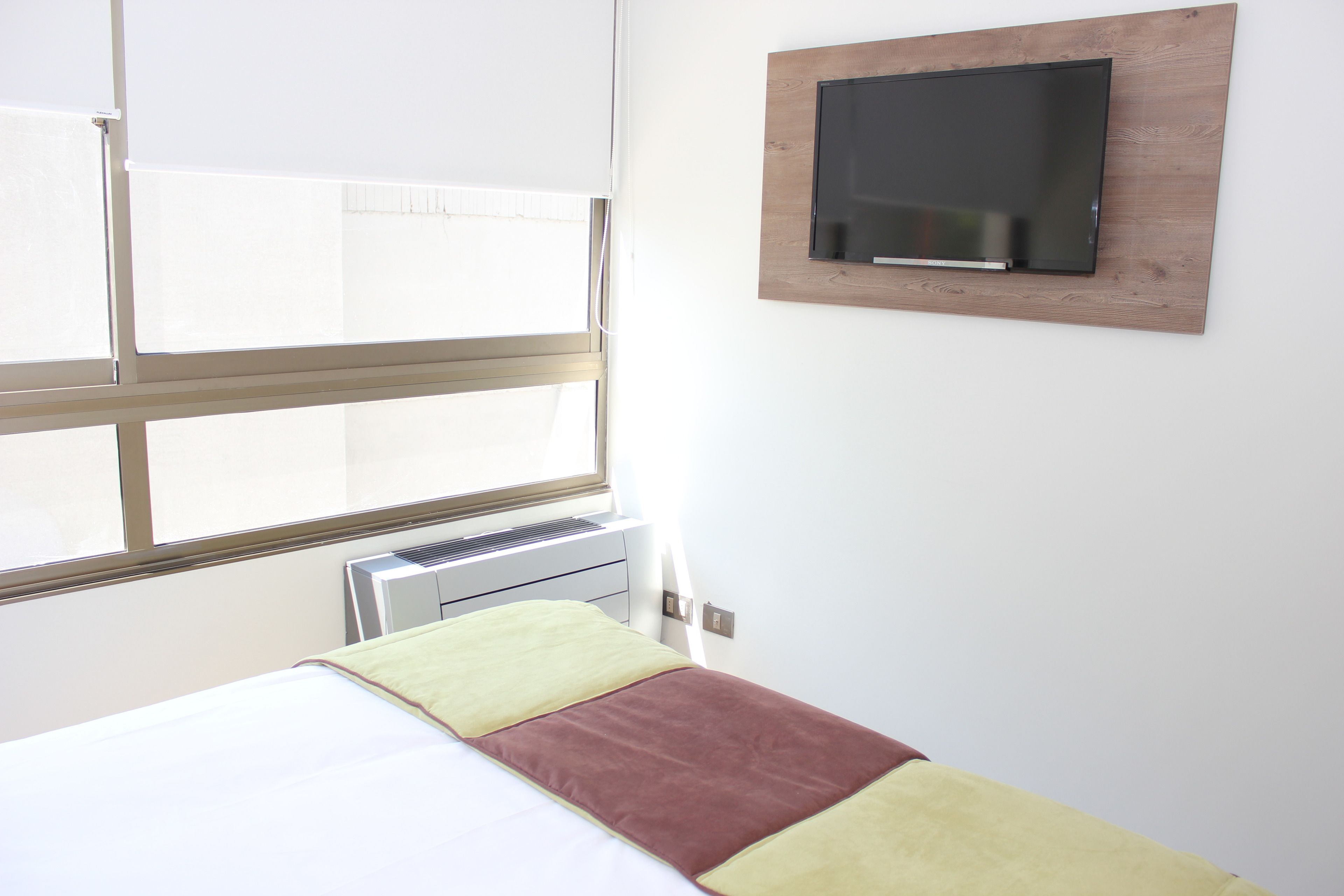 Una habitación de hotel con cama, ventana, aire acondicionado y televisor montado.