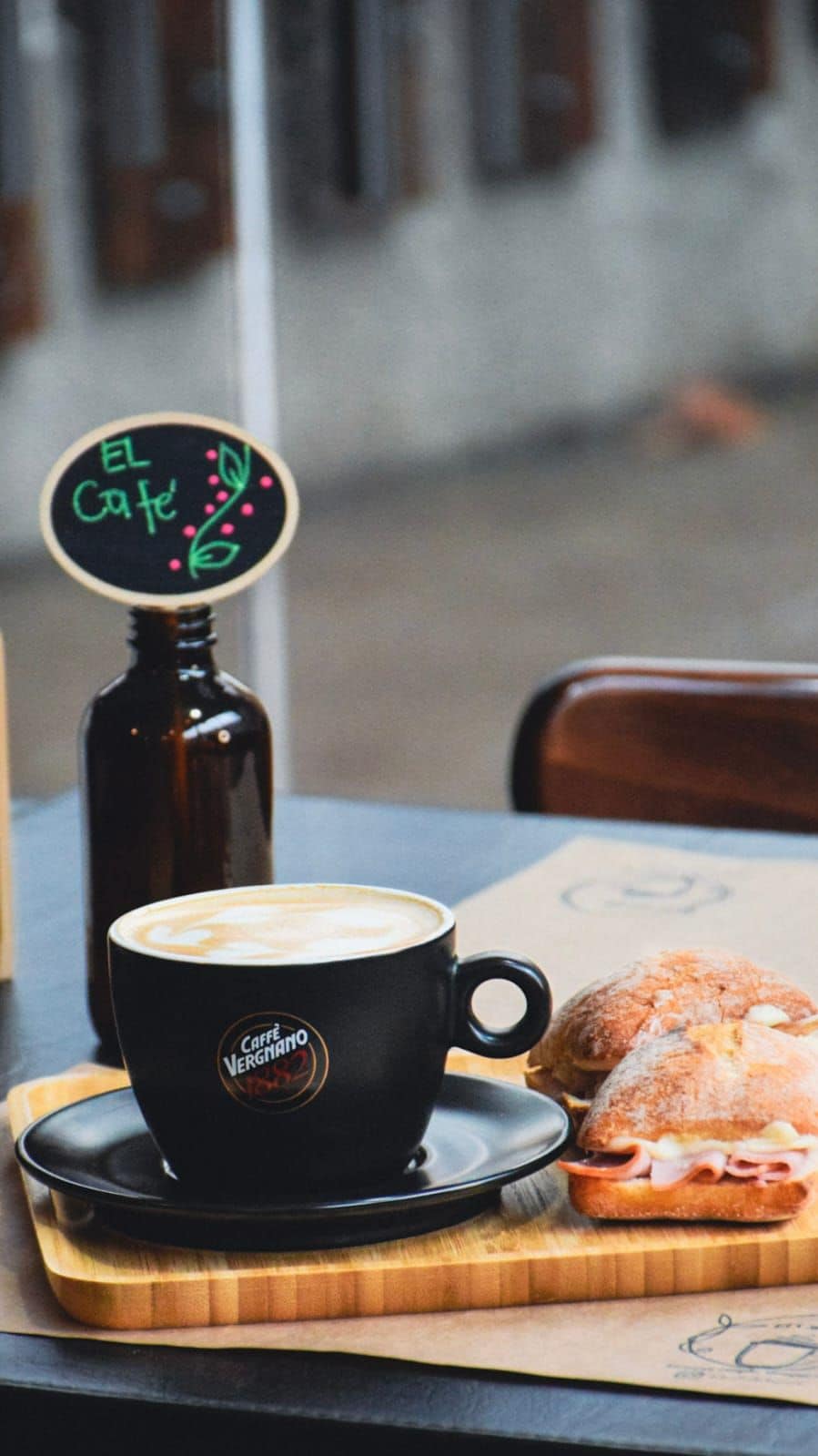 A cup of coffee with a logo on the saucer, a bottle, and a sandwich on a wooden board.