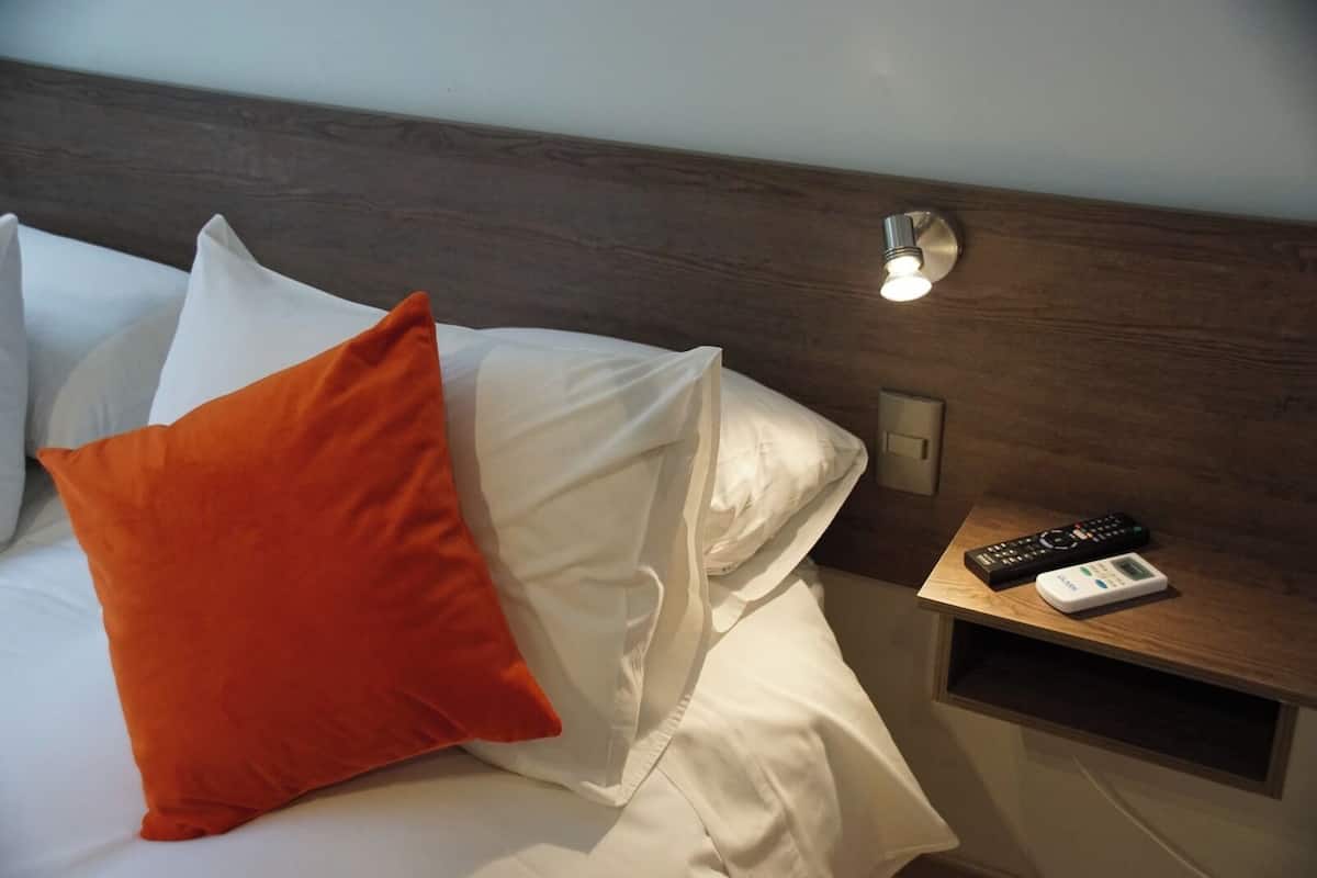 Una habitación de hotel con una cama bien hecha, una mesita de noche con un control remoto y una lámpara de pared.