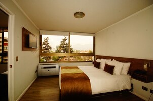 Habitación de hotel con una cama grande, una ventana con vista a la ciudad, un televisor montado y un escritorio pequeño con un teléfono.