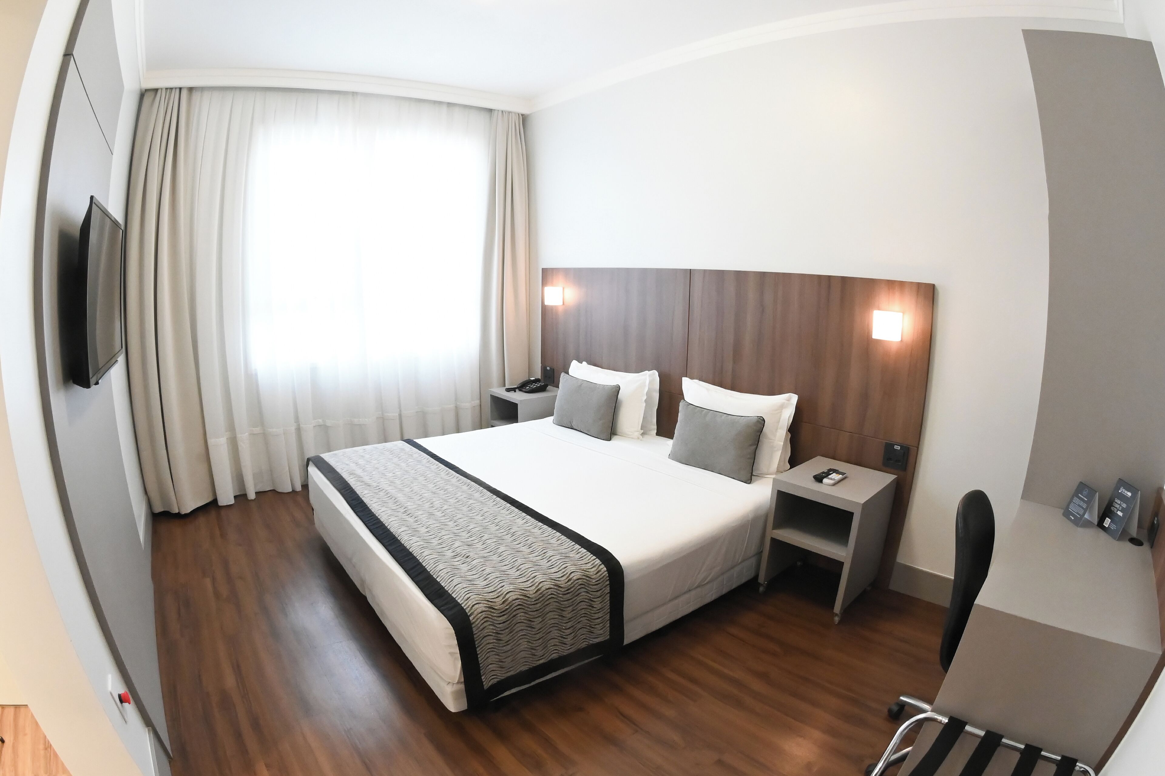 A hotel room with a large bed, a bedside table, a chair, a TV, and a window with curtains.