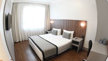 Habitación de hotel con una cama grande, una mesita de noche, una silla, un televisor y un ventanal con cortinas.