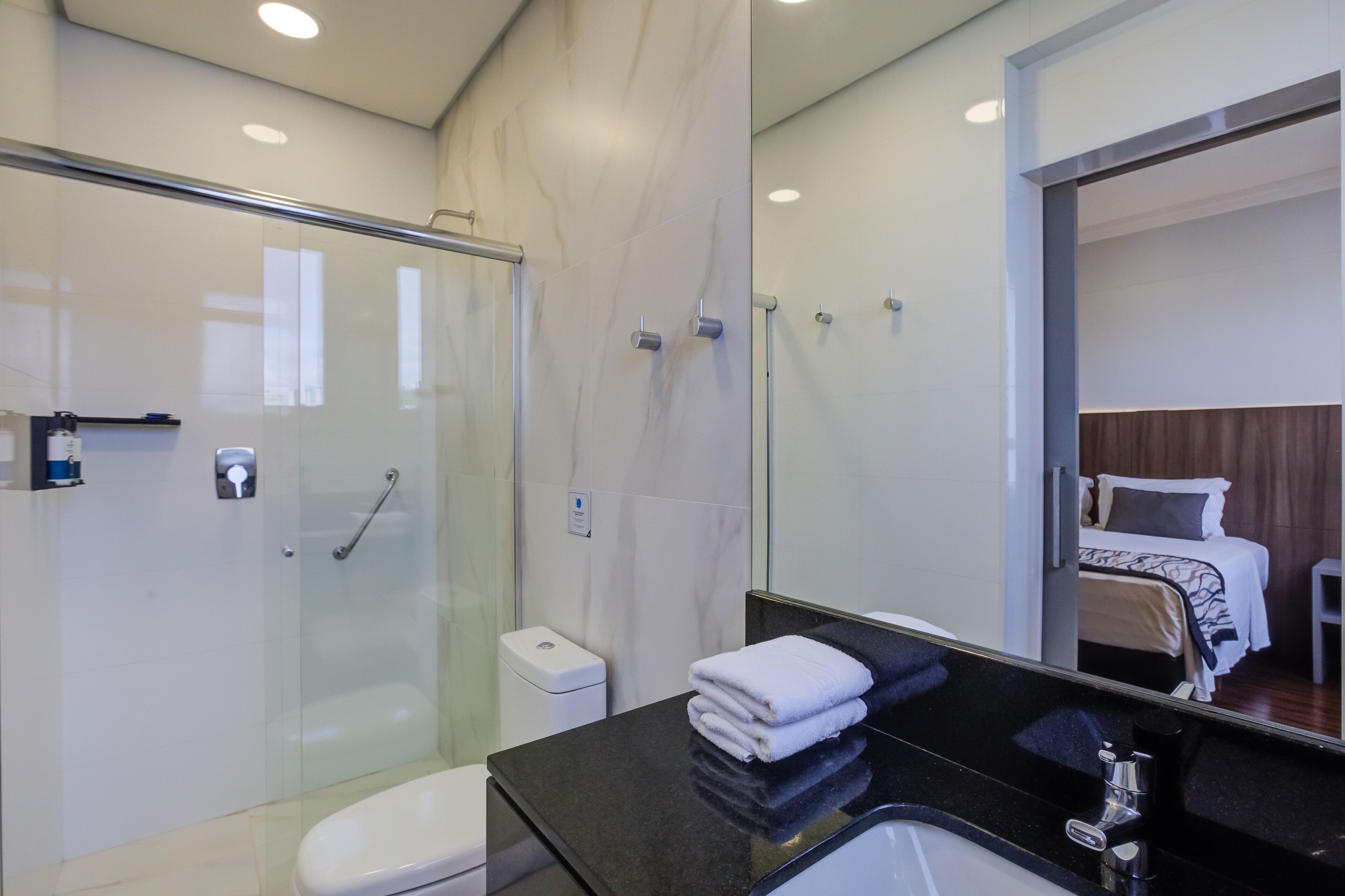 A modern bathroom with a glass shower enclosure, a black countertop, and a visible bed in the adjacent room.