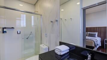 Un baño moderno con cabina de ducha de vidrio, encimera negra y una cama visible en la habitación contigua.