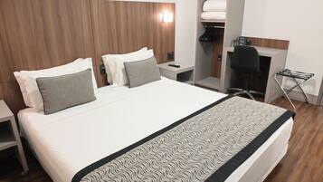 Quarto de hotel moderno com uma cama grande, escrivaninha, cadeira e roupeiro embutido.
