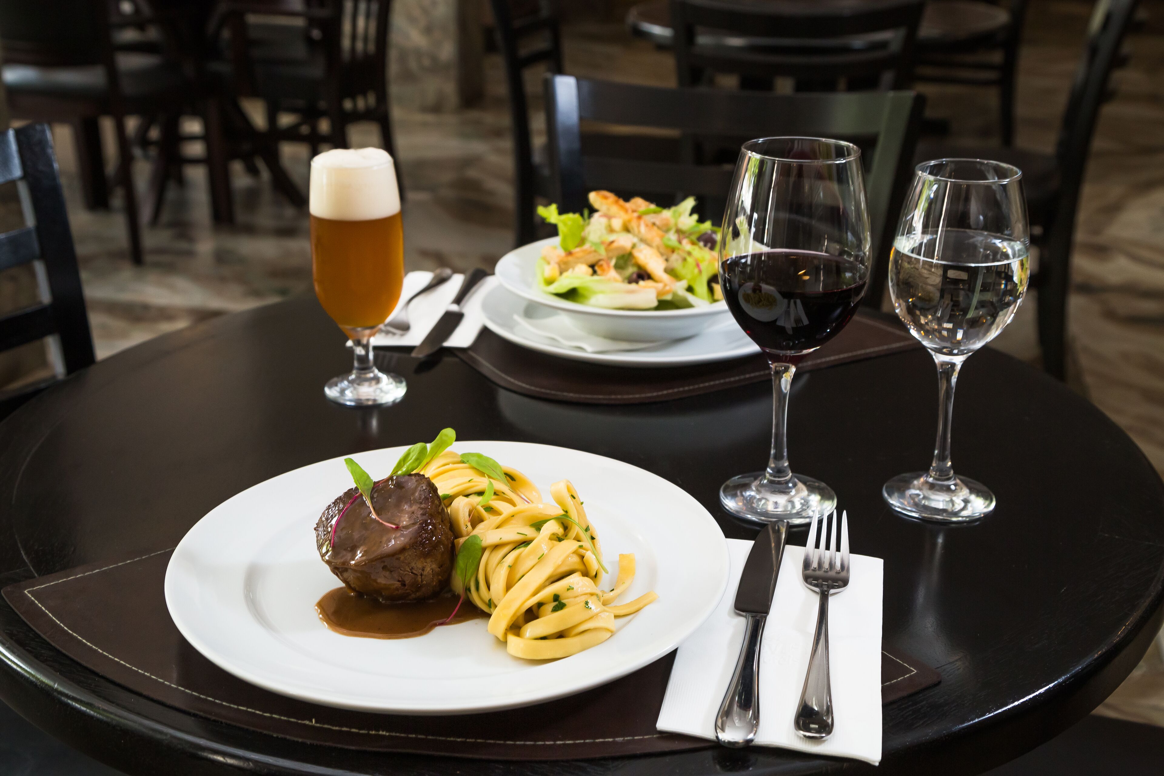 A dining table with a plate of pasta and meat, two glasses of wine, and a beer glass.