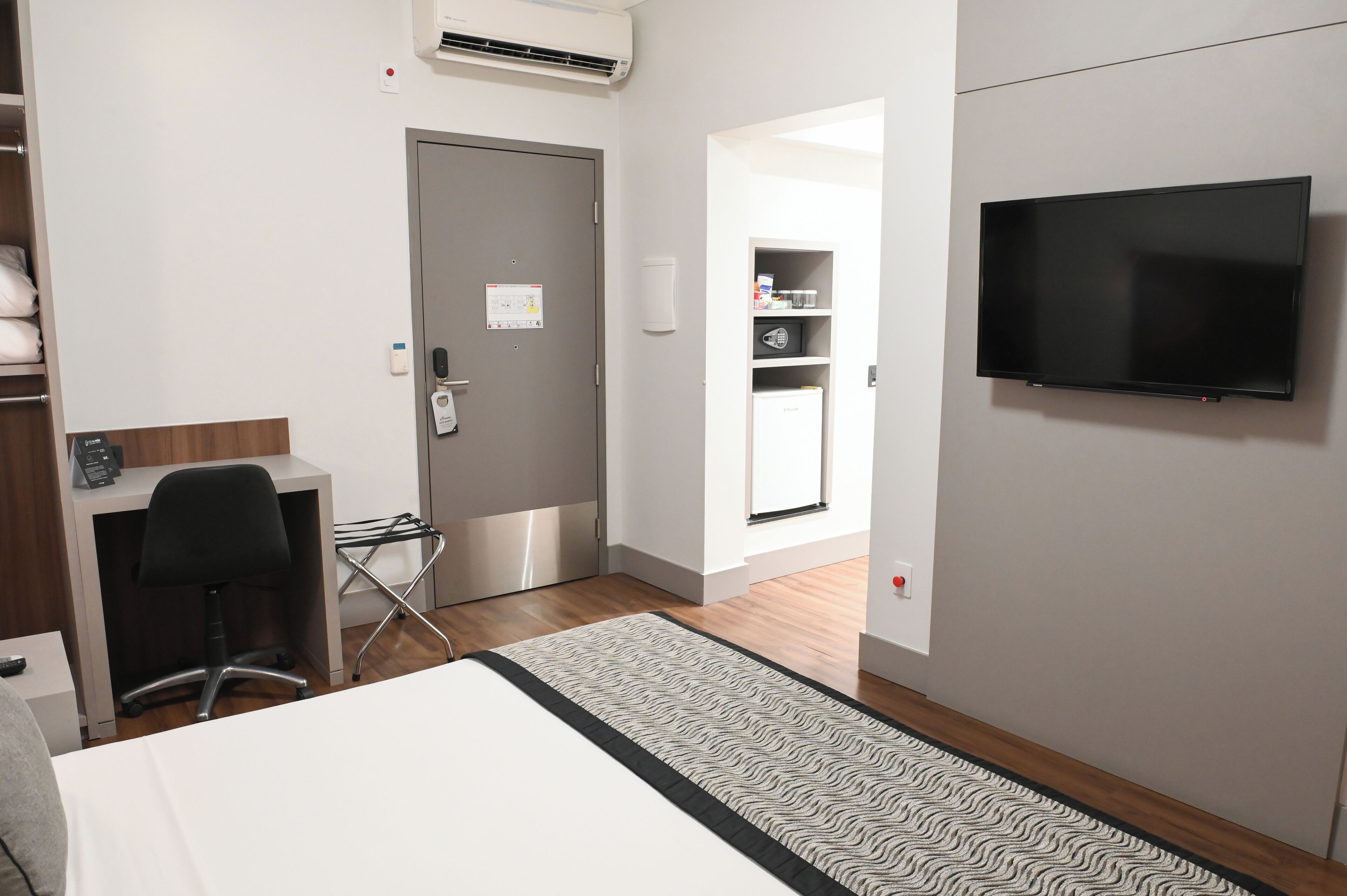 A hotel room with a bed, a desk with a chair, a TV mounted on the wall, a door, and a small kitchenette.