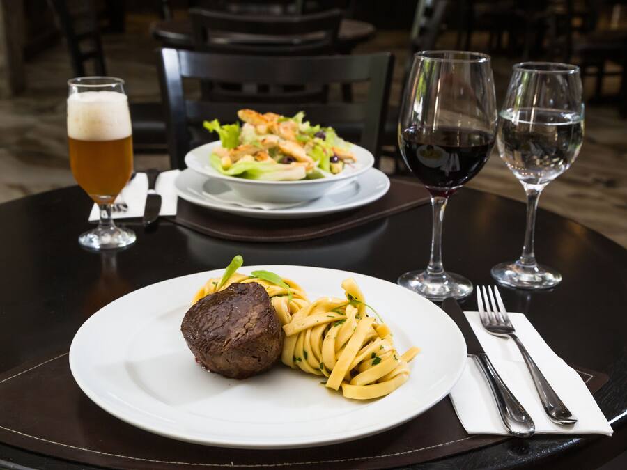 Una mesa de comedor con un plato de bistec y papas fritas, una cerveza en un vaso y dos copas de vino.