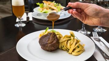 A plate of food with a steak and pasta, a glass of beer, and a spoon with sauce on a table.