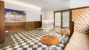 Una hall con pavimento a scacchi, una reception, un'area lounge con testiera capitonata e un'ampia finestra con vista sulla città.