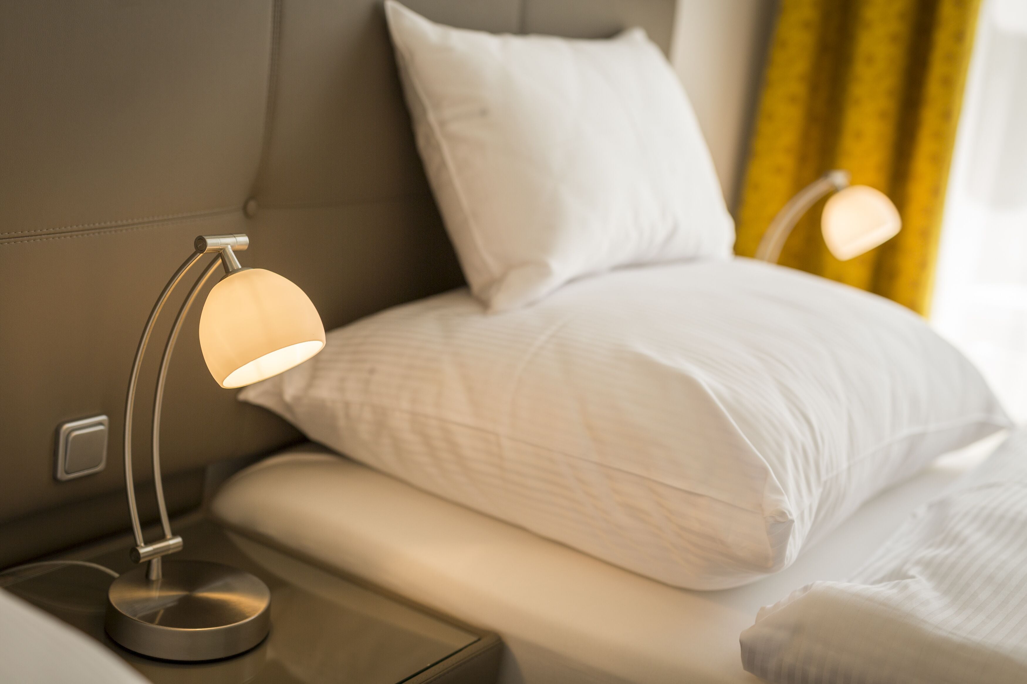 A neatly made bed with white linens and pillows, a bedside table with a lamp, and a headboard.