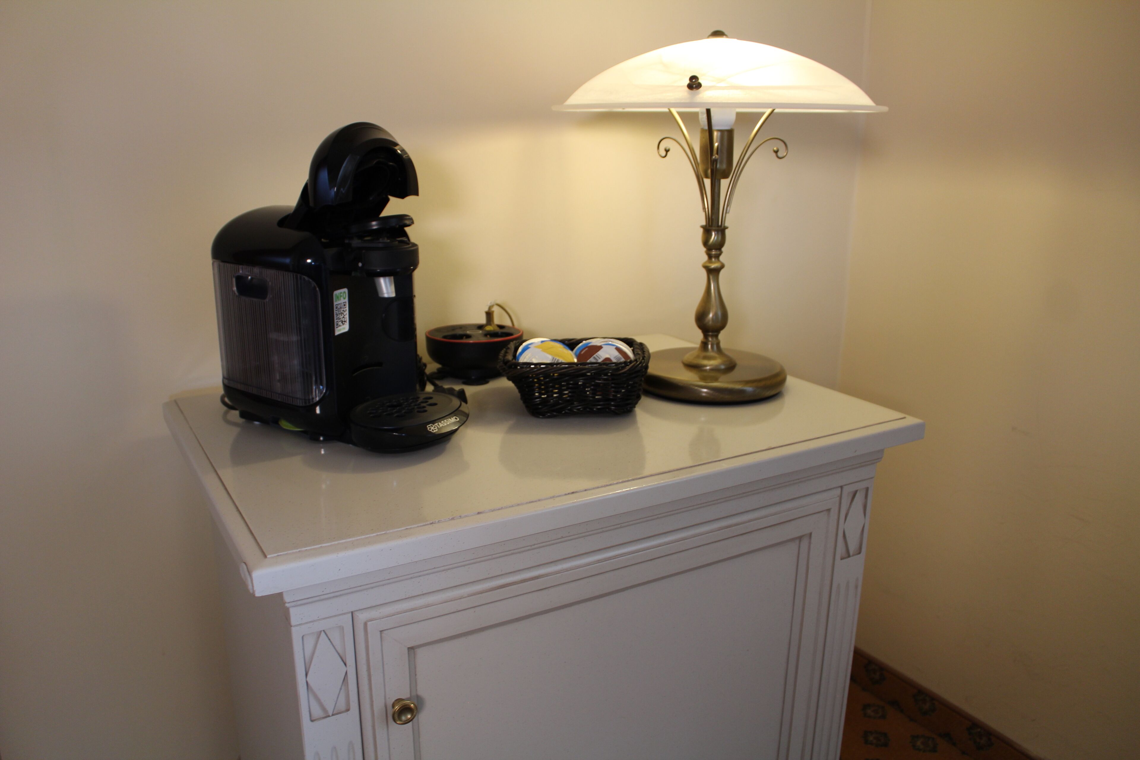 Una cafetera, una lámpara y una cesta con objetos sobre un mueble blanco.