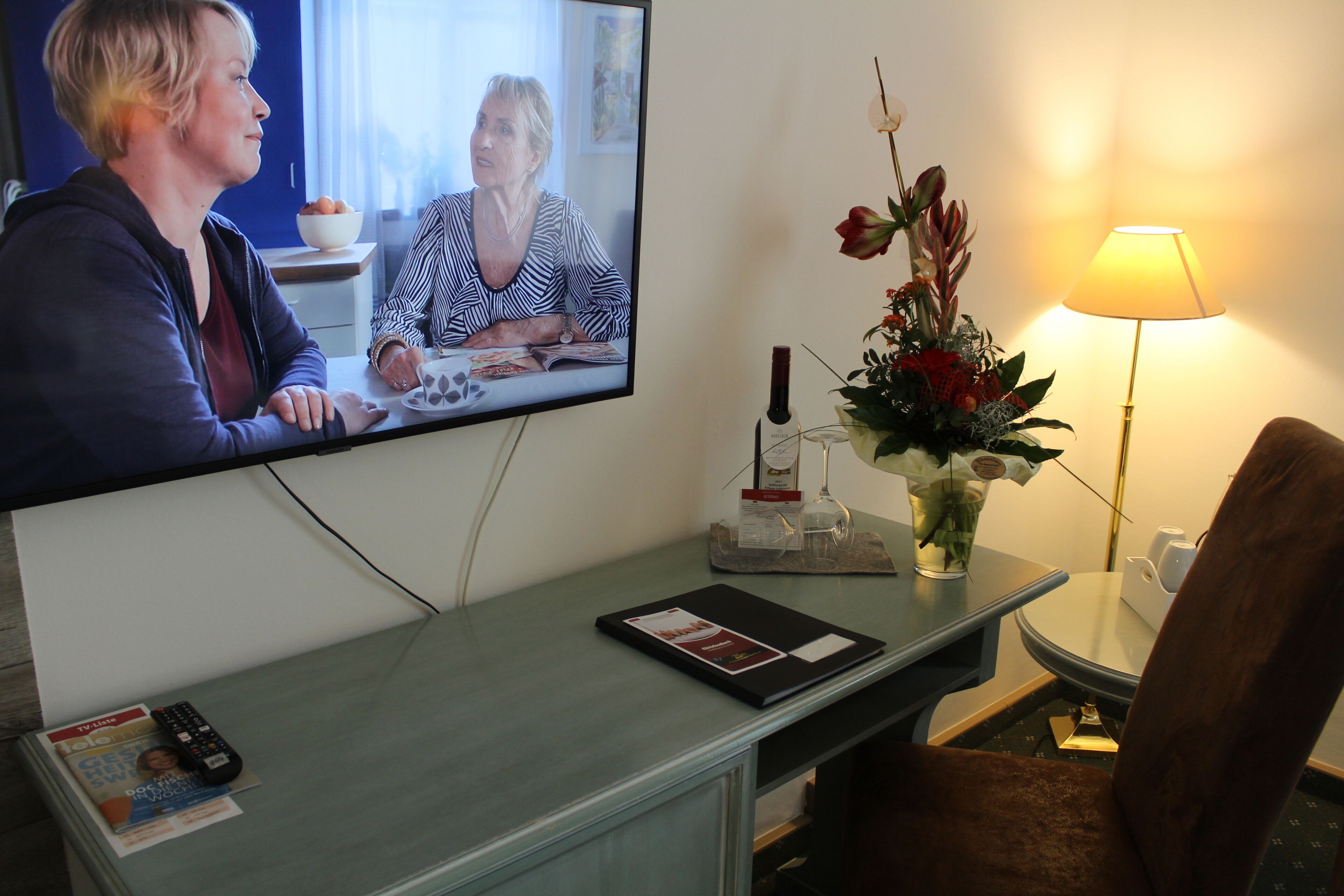 A hotel room with a television displaying a video call, a desk with a magazine, a remote control, a vase with flowers, and a lamp.