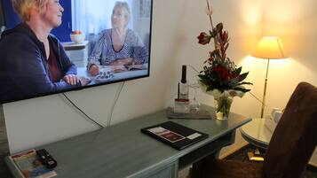 Habitación de hotel con una televisión mostrando una videollamada, un escritorio con una revista, un control remoto, un florero con flores y una lámpara.