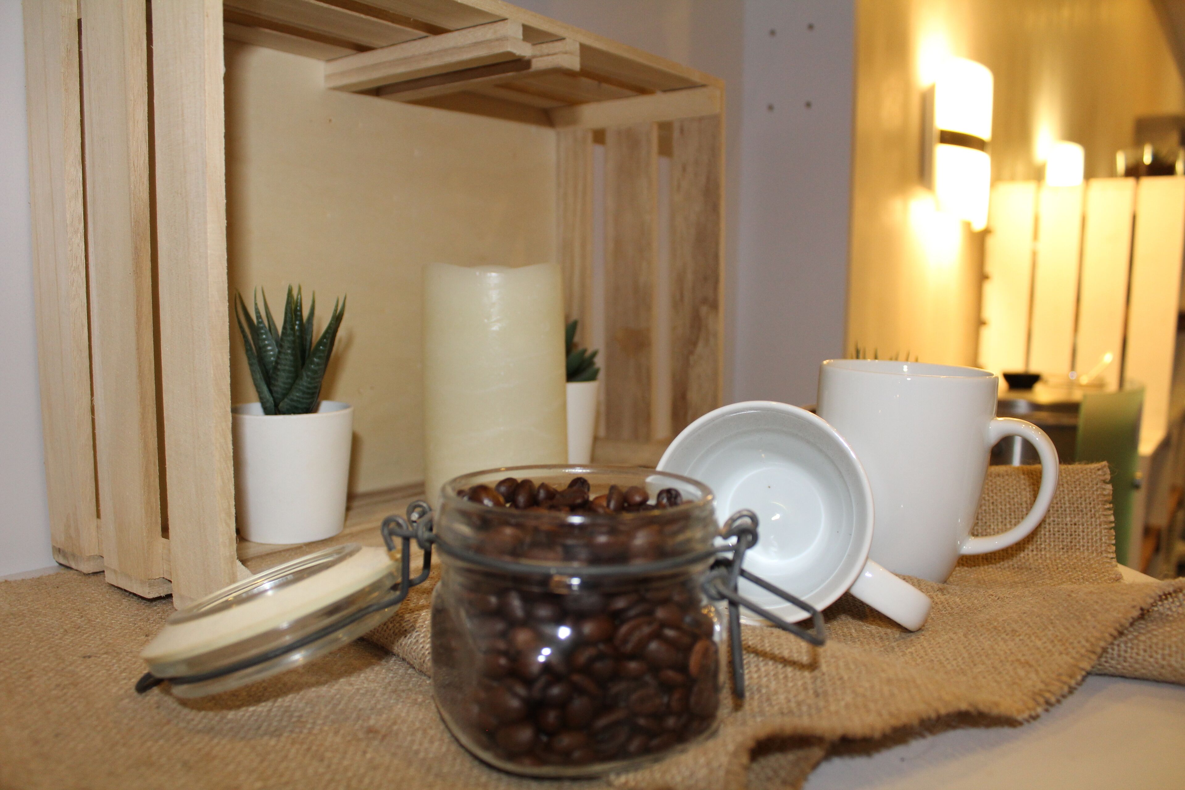 Una estantería de madera con un tarro de cristal de granos de café, una taza blanca y una pequeña planta en maceta.