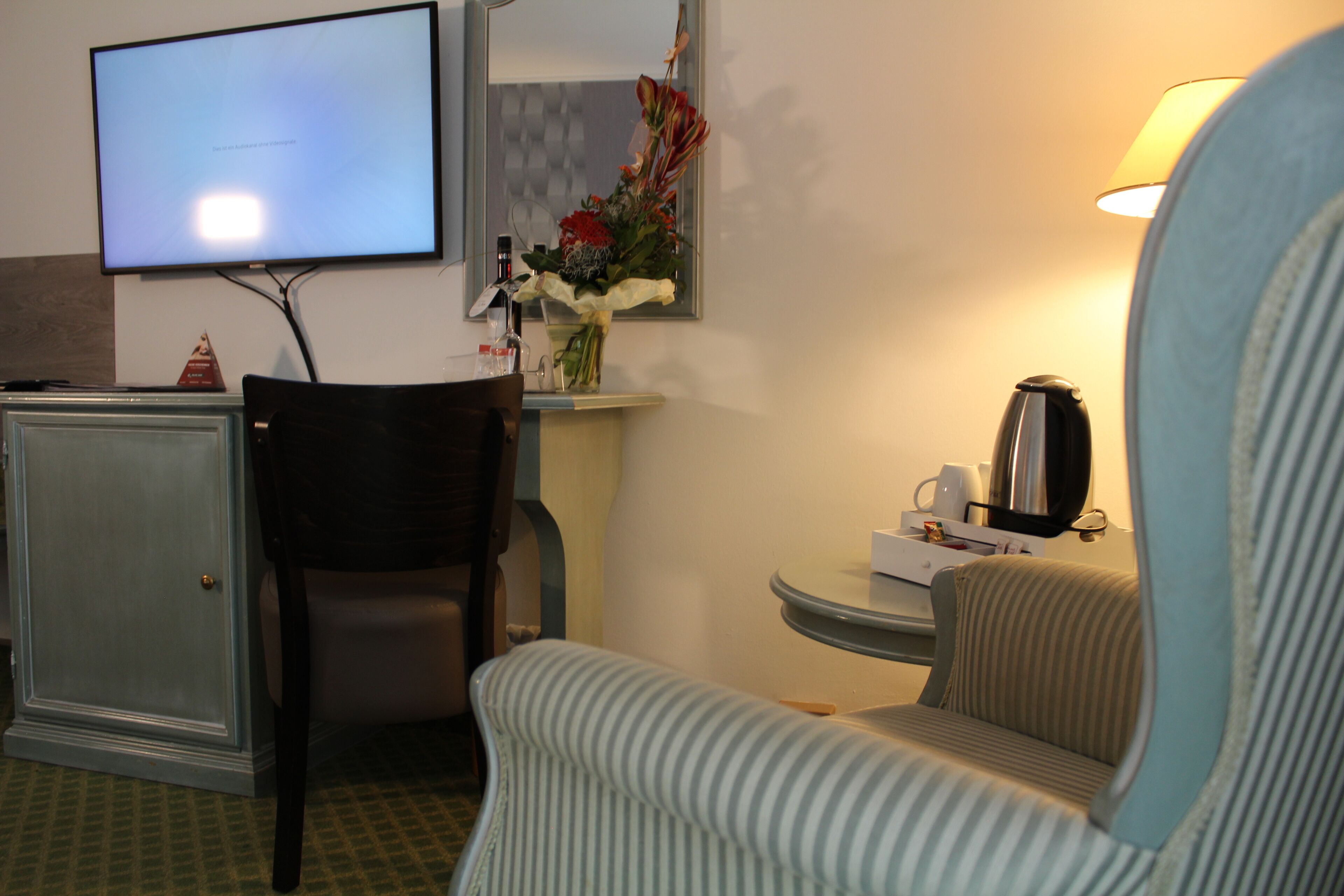 Ein Hotelzimmer mit an der Wand montiertem Fernseher, einem kleinen Tisch mit Lampe, einem Sessel und einem Schrank.