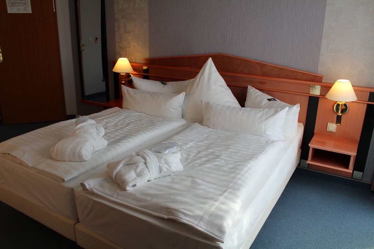 Een tweepersoonsbed met witte lakens en kussens, twee bedlampjes en een houten hoofdbord.