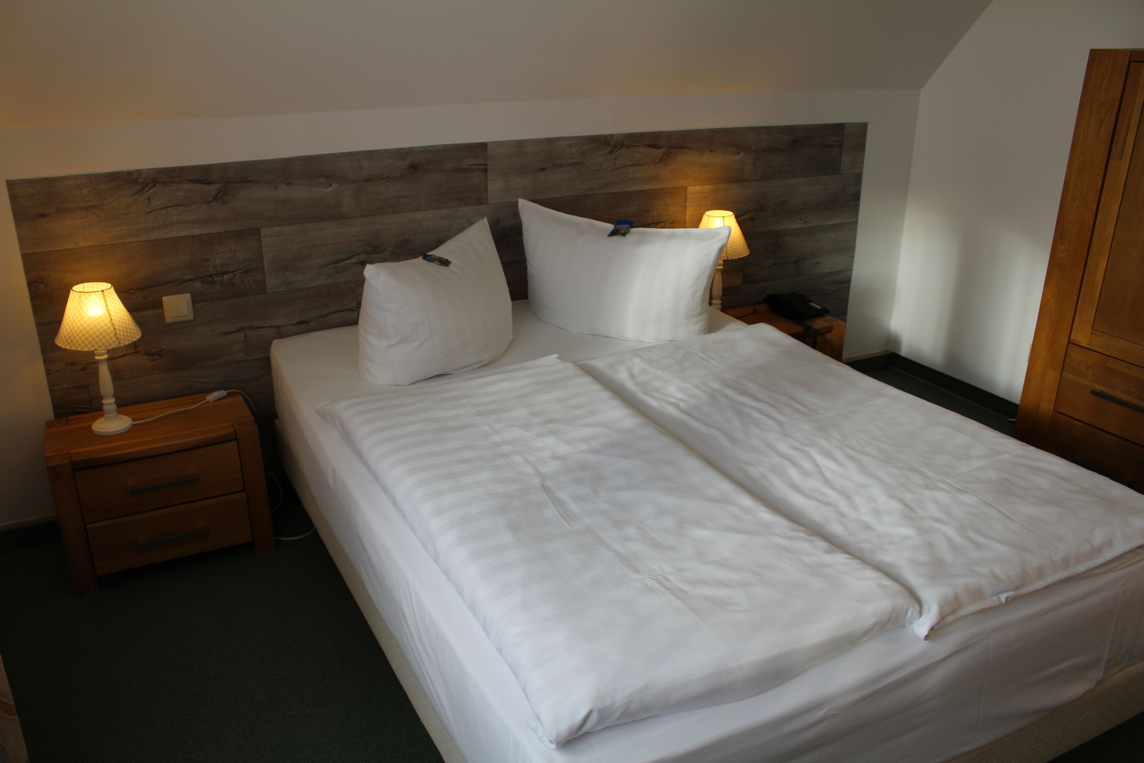 Een net opgemaakt bed met witte lakens, een houten hoofdbord en twee bedlampjes.