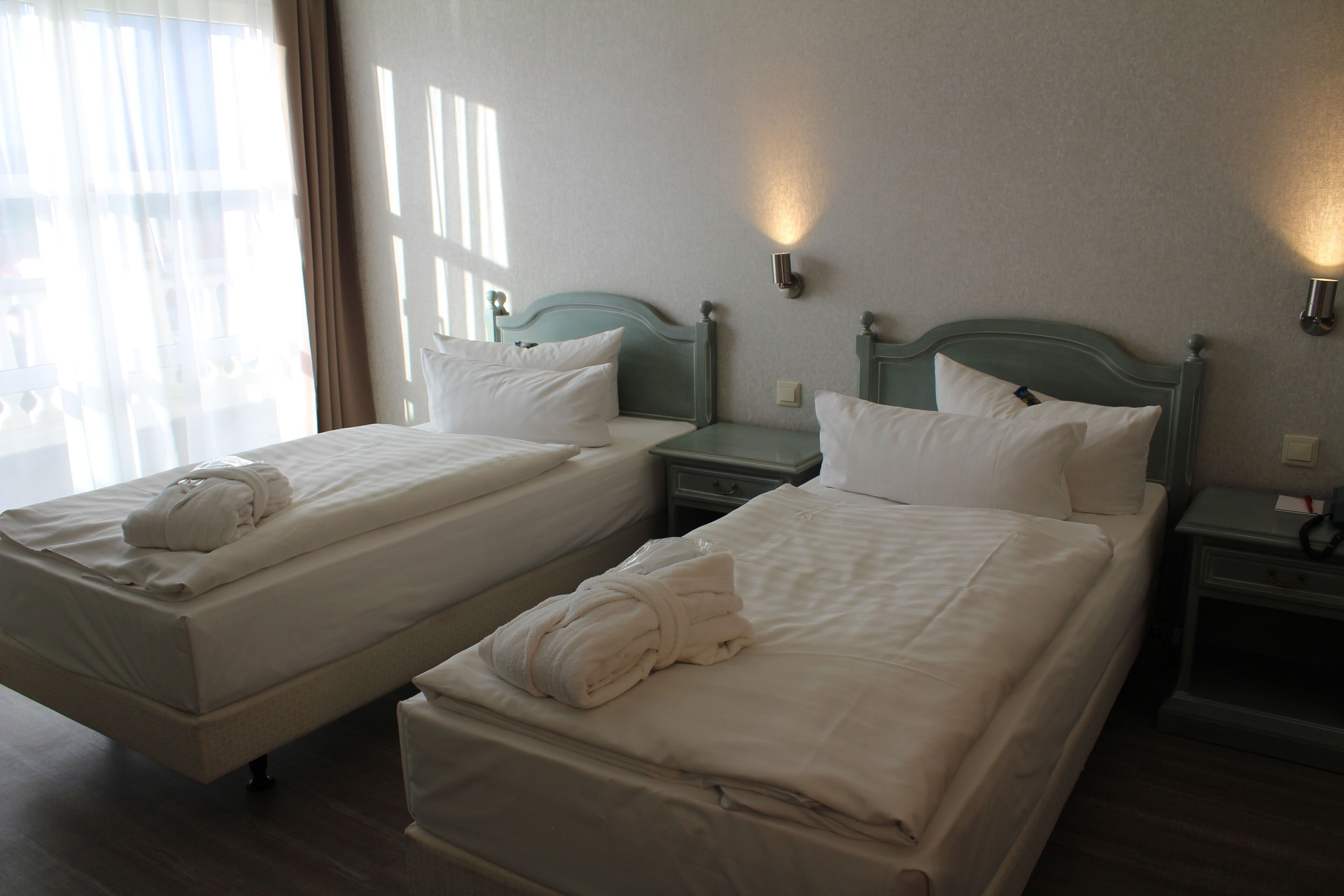 Two single beds with white linens and towels, wooden headboards, and bedside tables with wall-mounted lamps.