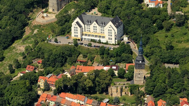 An aerial view of a historic building with a prominent tower, surrounded by lush greenery and smaller residential buildings.