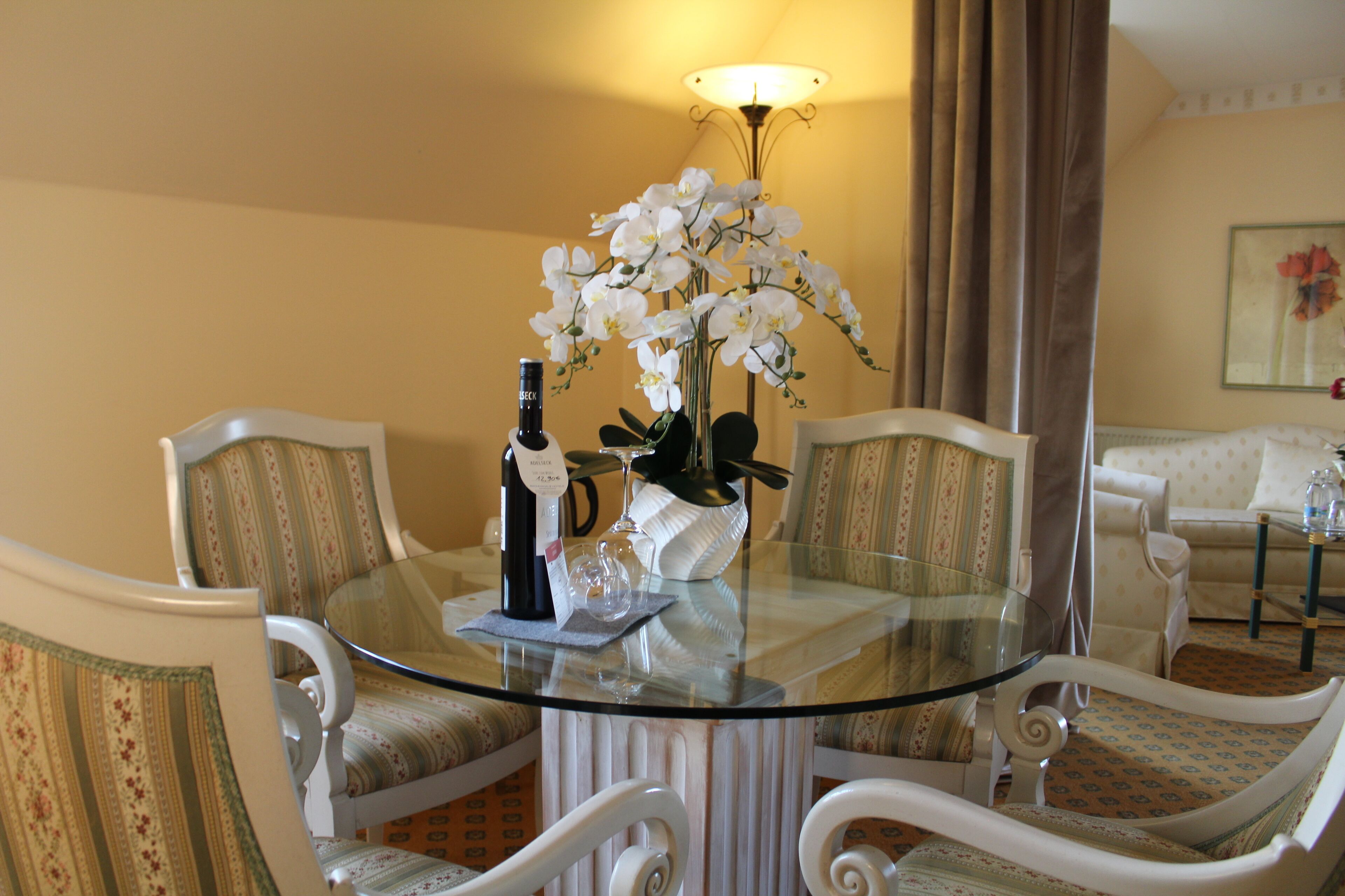 Uma área de refeições com uma mesa de vidro, quatro cadeiras estofadas, um vaso com flores brancas, uma garrafa e uma taça de vinho.