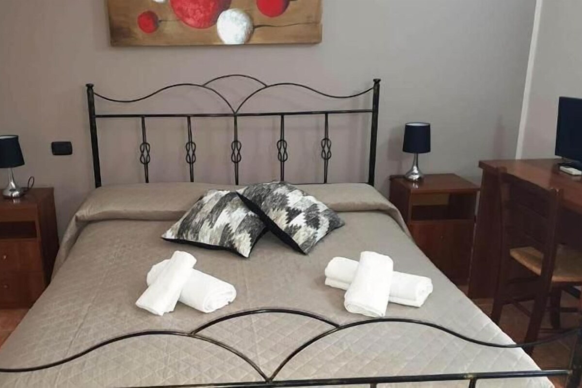 Un dormitorio ordenado con estructura de cama metálica, dos mesitas de noche con lámparas, un escritorio de madera y un cuadro en la pared.