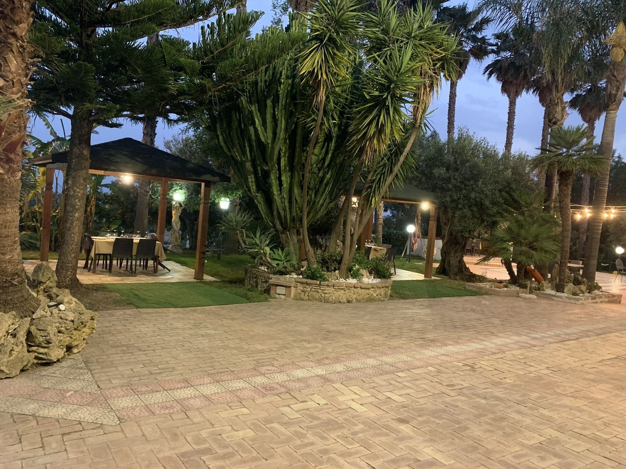 A paved outdoor area with a covered seating area, surrounded by palm trees and a statue.