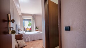 A hotel room with a bed, a bedside table, a nightstand, a window with curtains, a door, and a wall-mounted light switch.