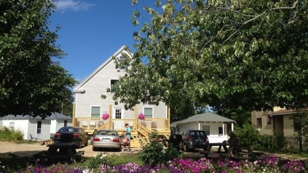 A two-story house with a front porch, a driveway with parked cars, and a flower bed with purple flowers. - Pine View Lodge (Old Orchard Beach)