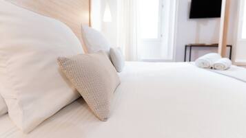 A neatly made bed with white linens and a textured pillow, a flat-screen TV mounted on the wall, and a window with curtains.
