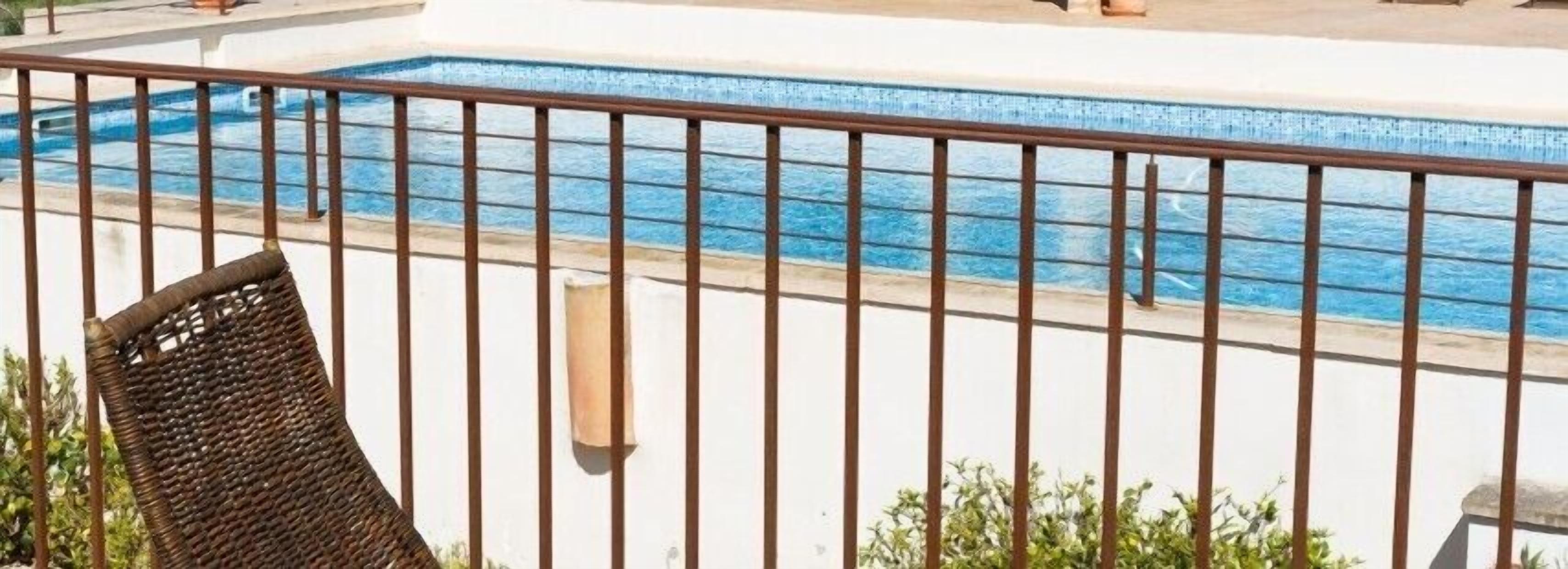 Una piscina con barandilla metálica, una silla de mimbre y algunas plantas en primer plano.