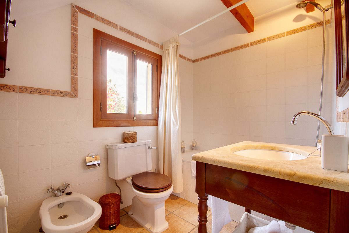 A bathroom with a toilet, bidet, sink, and a window with a curtain.