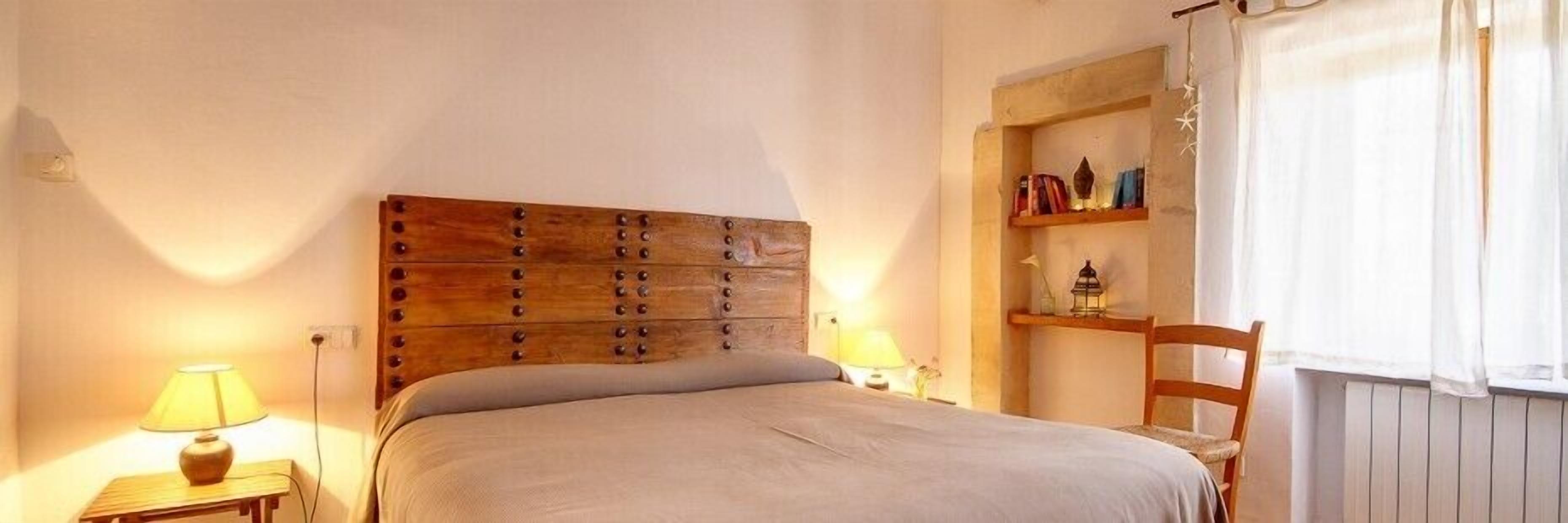 A bedroom with a wooden headboard, a bedside table with a lamp, a chair, and a shelf with books and decorative items.