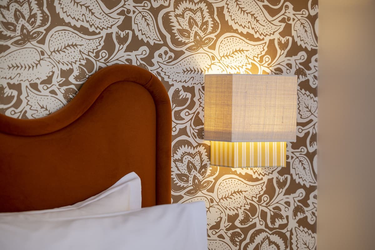 A bed with a brown headboard and white pillows against a patterned wallpaper.