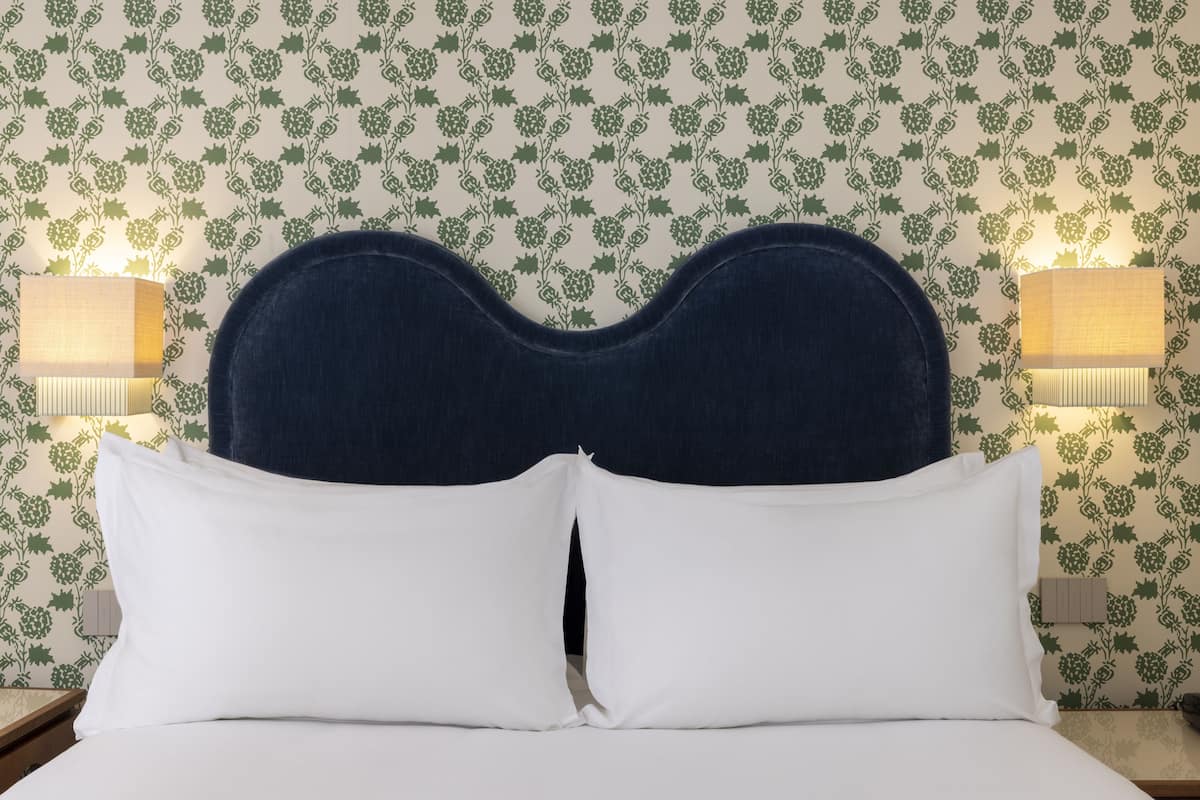 A bed with a dark headboard, white pillows, and a floral wallpaper background.