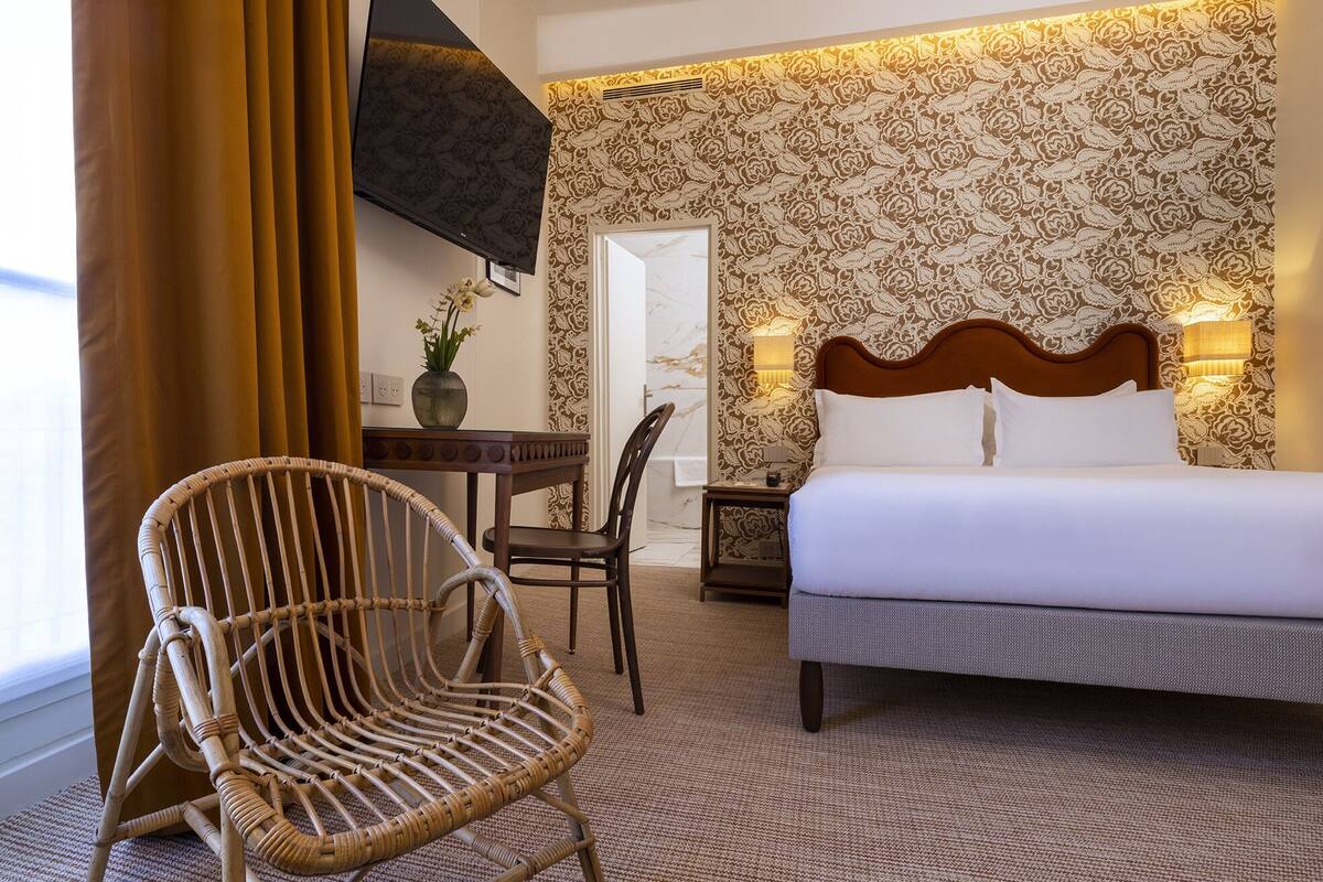 A hotel room with a bed, a wicker chair, a table with a vase, and a wall with a floral pattern.