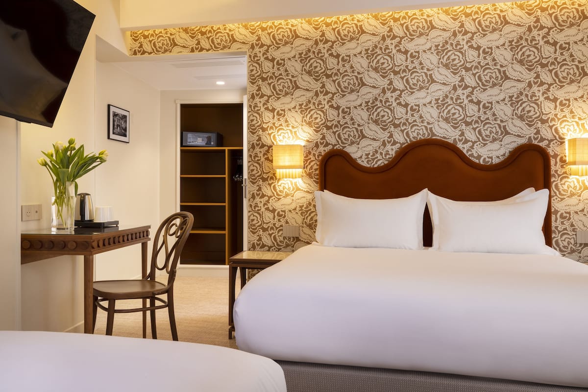 A hotel room with a large bed, a desk with a chair, a TV mounted on the wall, and a patterned wallpaper.