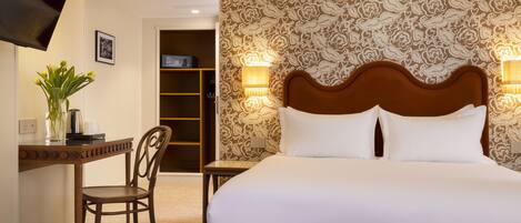 A hotel room with a large bed, a desk with a chair, a TV mounted on the wall, and a patterned wallpaper.