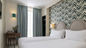 A hotel room with a large bed, a desk with a chair, a window with curtains, and a patterned wall. - Hôtel Michelet Odeon (Paris)