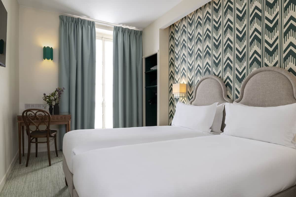 A hotel room with a large bed, a desk with a chair, a window with curtains, and a patterned wall.